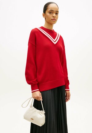 Kobieta ubrana w czerwony sweter z białymi paskami na kołnierzyku i mankietach, trzymająca małą białą torebkę, w połączeniu z czarną plisowaną spódnicą.