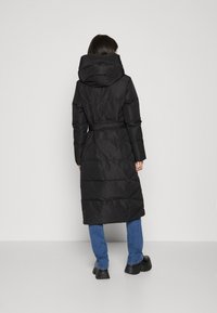 Kvinna som bär en lång svart vadderad kappa med stor huva, blå jeans och svarta platåskor, stående mot en enkel vit vägg.