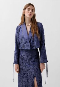 Kurze marineblaue Jacke mit strukturiertem floralen Muster, langen Ärmeln, Knopfdetails, getragen über einem passenden, taillierten Rock mit einem seitlichen Schlitz.
