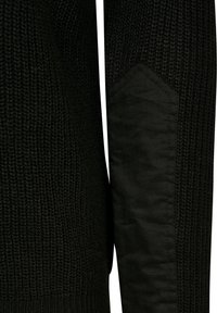 Manche de pull en tricot noir avec un renfort en tissu au niveau du coude, cousu en forme hexagonale.