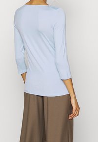Donna che indossa una maglietta azzurro chiaro a maniche tre quarti e pantaloni marroni larghi, vista di spalle su uno sfondo neutro.