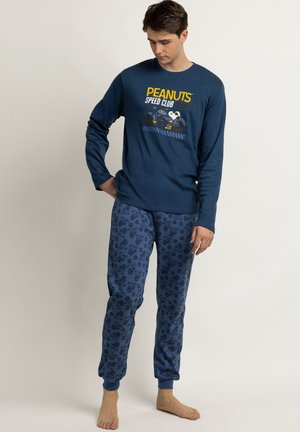 Joven con una camiseta de manga larga azul marino de Peanuts Speed Club y pantalones de pijama azules con estampado, de pie descalzo frente a un fondo liso.