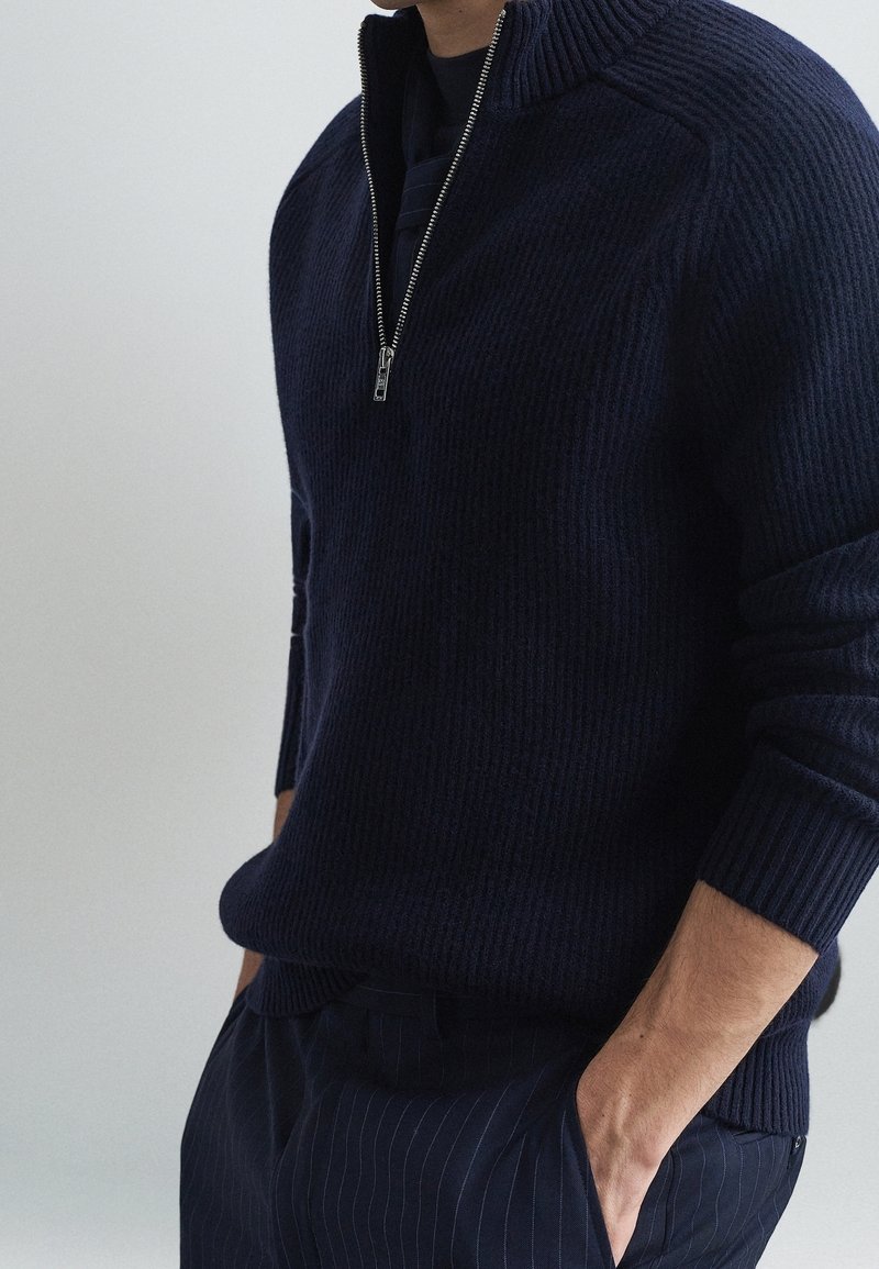 Homme portant un pull en maille côtelée bleu marine foncé avec une fermeture éclair demi-longueur à l'avant et les mains dans les poches d'un pantalon bleu marine à fines rayures.