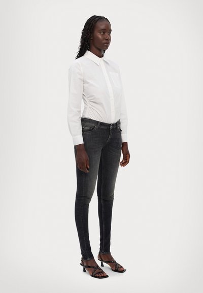 Hvit skjorte med knapper og brystlomme, lagt sammen med mørkegrå trange jeans og svarte sandaler med stropper og hæl, stående mot en nøytral bakgrunn.