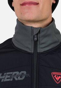 Svart jacka med grå detaljer, hög krage, dragkedjestängning, har en framträdande logotyp "HERO" och ett rött "ROSSIGNOL"-emblem på bröstet.