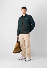 Donkergroene vest met ritssluiting en een eenvoudig ontwerp. Beigekleurige broek. Bruine tas met donkere accenten. Witte en groene sneakers.