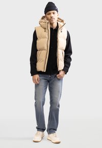Chaleco acolchado en beige con capucha, sobre una camisa negra con botones. Jeans ajustados en azul, combinados con zapatillas ligeras beige. Corte recto.
