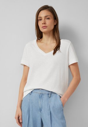 Vrouw met stijl haar en bruin haar die een wit T-shirt met V-hals en korte mouwen draagt en lichtblauwe high-waisted jeans, met één hand in de zak.