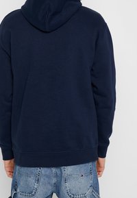 Persona con una sudadera azul marino y jeans de mezclilla azul claros, vista desde atrás contra un fondo claro.