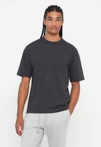 Νεαρός άνδρας με πλεξίδες, φορώντας ένα απλό γκρι κοντομάνικο μπλουζάκι και ανοιχτόχρωμες φόρμες, στέκεται με το ένα χέρι στην τσέπη.