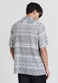 Chemise à manches courtes présentant un motif géométrique noir et blanc, avec un col pointu et une coupe décontractée. Le tissu semble léger et texturé.