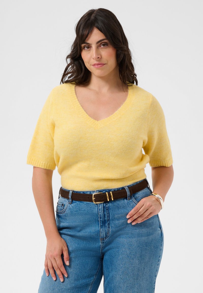Femme aux cheveux foncés portant un pull jaune à manches courtes et en col en V, rentré dans un jean bleu taille haute avec une ceinture marron sur fond blanc.