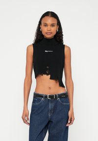 Fekete ujjatlan crop top álló nyakkal, egyenetlen szegéllyel és kopott élekkel; fehér logóval a mellrészen. Kék farmerral és fekete övvel párosítva.