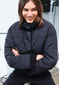 Giubbotto imbottito nero con collo alto, dotato di bottoni a pressione e texture morbida ai polsini. Tessuto liscio con design trapuntato.