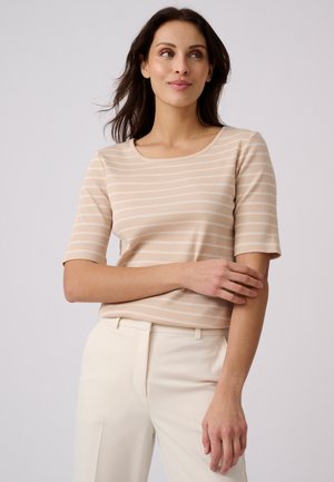 Vrouw die een beige-wit gestreept shirt met korte mouwen en crèmekleurige broek draagt, kijkt opzij met één arm over haar lichaam gekruist.