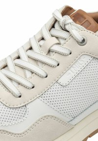 Sneaker beige e bianco con pannello laterale testurizzato, spesse stringhe bianche legate a fiocco, dettagli in suede e una linguetta in pelle marrone.