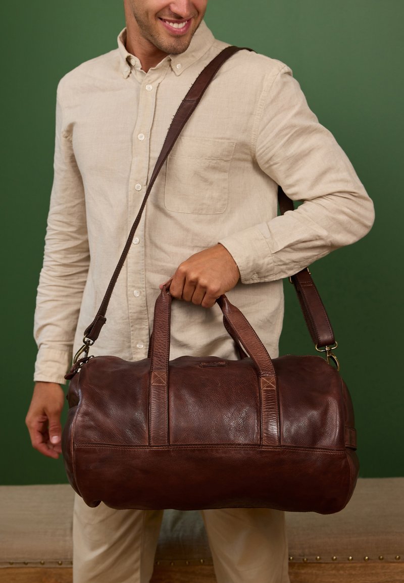 Borsa da viaggio in pelle marrone con doppi manici e una tracolla staccabile, caratterizzata da una texture liscia e dettagli di cucitura sottili.