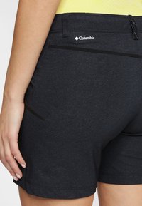 Närbild av en person som bär svarta Columbia-shorts med blixtlås på bak- och sidofickorna, och en delvis vy av deras vänstra hand som vilar vid sidan.