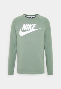 Zelená mikina vyrobená z měkké tkaniny, která má velké bílé logo Nike přes hrudník a raglánové rukávy pro uvolněný fitting.
