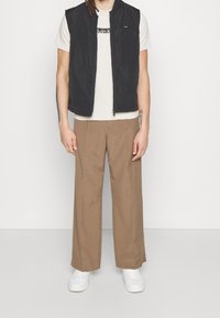 Černá bezrukávová vesta přes světle béžové tričko, kombinovaná s hnědými širokými kalhotami a bílými teniskami. Hladké textury a jednoduchý design.