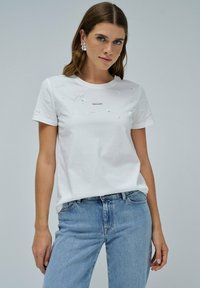 Camiseta blanca de algodón con bordado floral y un pequeño logo plateado en el frente. Cuello redondo y mangas cortas combinadas con unos vaqueros azul claro.