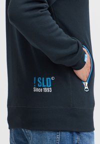 Mørkeblå sweatshirt med lynlåslomme og et hvidt og lyseblåt logo "SLD Since 1993" på forsiden. Glat tekstur.