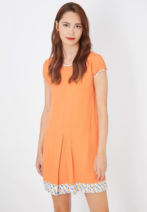Joven mujer con cabello largo castaño, vestida con un vestido naranja de manga corta con un borde blanco decorado en las mangas y el dobladillo, de pie contra un fondo blanco.