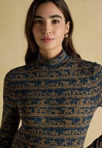 Długi rękaw turtleneck w granatowym kolorze, z złotymi liniowymi rysunkami koni i postaci w układzie siatki. Gładka tekstura i dopasowany krój.