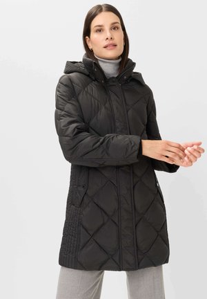 Woman wearing a black quilted hooded coat and gray turtleneck, adjusting her hands against a plain light background.
