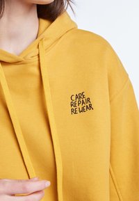 Sudadera amarilla hecha de un tejido suave con un bolsillo canguro y capucha ajustable con cordones. Texto "CUIDADO REPARAR RE:USAR" bordado en negro.