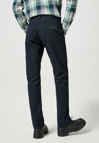Marinblå chinos i bomull med en slät textur, som har två bakfickor och en läderlogopatch vid midjan. Svarta skor synliga.