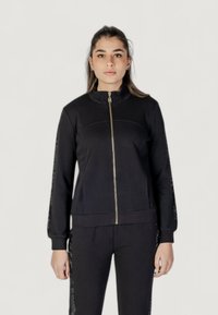 Giacca nera con zip e colletto alto, caratterizzata da maniche affusolate e dettagli testurizzati lungo il lato, realizzata in tessuto morbido.