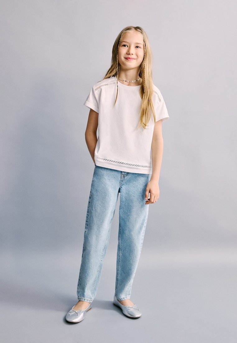 Mlada djevojka stoji, nosi bijelu košulju kratkih rukava, svijetloplave traperice, srebrne baletanke i ogrlicu od perlica na jednostavnoj sivoj pozadini.