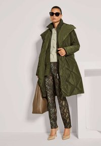 Olijfgroene gewatteerde lange jas over een licht trui, gecombineerd met slangenprintbroek en beige hakken. Geaccentueerd met zwarte zonnebril en een bruine tas.