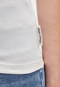 Άσπρη ριμπ λινάτσα με κυρτή τελειώματα, με μια μικρή ετικέτα υφάσματος που γράφε�ι "Marc O'Polo" στο πλευρικό ραφή. Φορεμένη με μπλε τζιν.