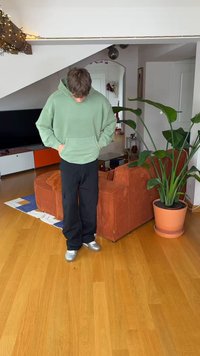 Zelená mikina s kapucí a přední kapsou, černé volné kalhoty a stříbrné boty. Vnitřní prostředí s rostlinou a oranžovou texturovanou pohovkou.