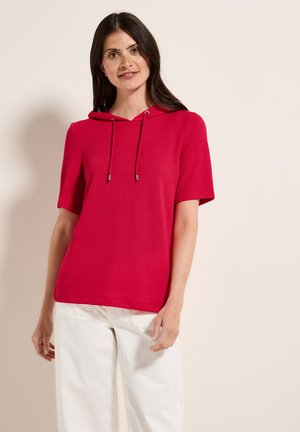 Hoodie rouge à manches courtes en tissu tricoté texturé, avec un cordon de serrage au cou et une coupe décontractée. Associé à un pantalon large blanc.