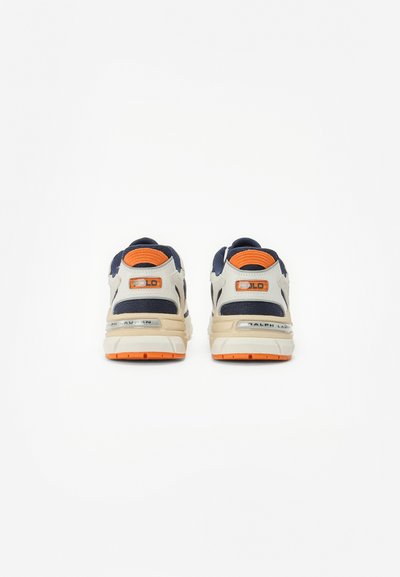 Sapatilhas beige e azul-marinho com detalhes em laranja, apresentando um superior em tecido texturizado, sola acolchoada e detalhe da marca no calcanhar.