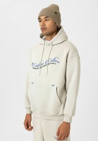 Lichtbeige hoodie met een voorzak, trekkoord en blauwe grafische tekst. Draagt met een lichtbruine gebreide beanie en bijpassende broek. Zachte textuur.