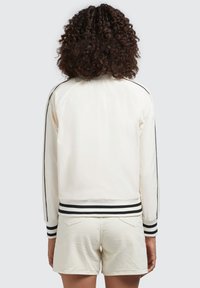 Vit bomberjacka med svarta randiga muddar och nederkant, med en slät textur och raglanärmar, som matchas med ljusa shorts.