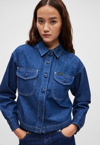 Sötétkék farmerdzseki, gombos elülső résszel, két mellzsebbel, varrott részletekkel és hosszú ujjal, gombos mandzsettával.