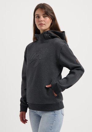 Vrouw met lang bruin haar, gekleed in een donkergrijze hoodie met een subtiel logo en lichtblauwe jeans, hand in de zak.