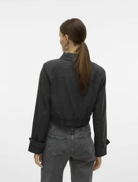 Lühendatud, tumeda halli denimpakk, millel on sobitatud vöökoht, nööpidega mansetid ja sile tekstuur. Tagantvaates on näha minimaalsed õmbluse detailid.