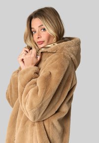 Veste à capuche en fausse fourrure beige, présentant une texture douce, un design surdimensionné et une grande fermeture éclair. Un tissu lisse enveloppe confortablement le porteur.