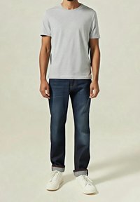 Homme portant un t-shirt gris clair à manches courtes, un jean bleu foncé retroussé et des baskets blanches, debout devant un fond uni.