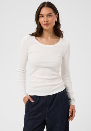Femme aux cheveux foncés portant un haut blanc à manches longues et un pantalon foncé, debout avec une main dans la poche sur un fond uni clair.