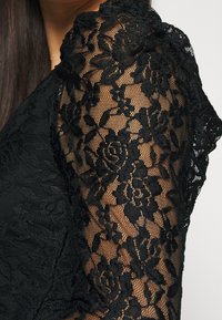 Schwarzes Spitzenoberteil mit floralen Mustern, durchscheinender Textur und figurbetontem Design, mit leicht gepufftem Ärmel und wellenförmigen Kanten.