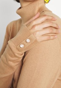 Maglione beige a collo alto con polsini a costine, caratterizzato da due bottoni rotondi simili a perle. Tessuto morbido con silhouette aderente.