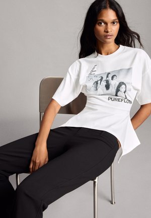 Femme aux longs cheveux foncés assise sur une chaise, portant un t-shirt blanc avec un motif graphique et un pantalon noir sur un fond uni gris clair.