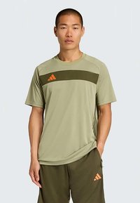 Camiseta atlética verde oliva hecha de una tela suave, con un panel de oliva oscuro y rayas naranjas en el pecho. Mangas cortas, ajuste relajado.